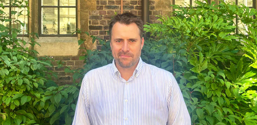 Tom Worthington, smiling, in blue shirt with hedges in the background